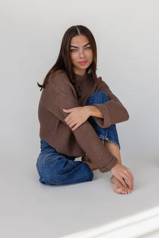 Woman wearing a brown sweater and blue jeans sitting on a white floor.