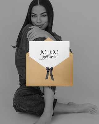 Woman holding a JO+CO gift card with a brown envelope and black bow on a gray background