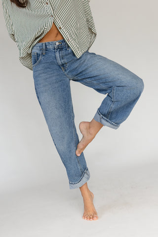 A person wearing light blue straight-leg denim jeans with rolled cuffs, paired with a green and white striped shirt, shown from the waist down against a plain light gray studio background.