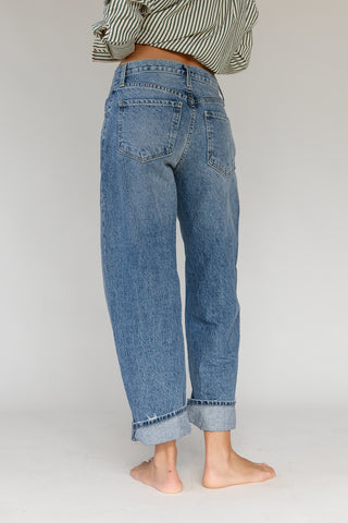 A person shown from the back wearing light blue relaxed-fit denim jeans with rolled cuffs, standing barefoot against a plain light gray studio background.